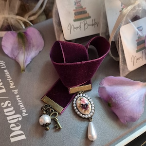 May include: A deep purple velvet ribbon bookmark featuring a brass-toned metal clasp, a small antique-style key charm, a round white faux pearl bead, and an oval cameo pendant with a floral rose design surrounded by tiny white faux pearls.
