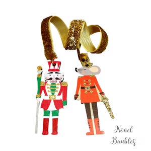 Op de afbeelding: Twee kerstversieringen: een notenkrakersoldaat en een muizensoldaat, beide met gouden glitterlinten. De notenkraker is rood, wit en groen. De muis draagt een oranje uniform. De tekst "Novel Baubles" staat rechtsonder.