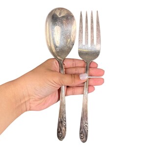 May include: A large silver serving spoon and fork set. The spoon has a rounded bowl, and the fork has four tines. Both utensils have ornate floral designs on the handles. The set is held against a white background.