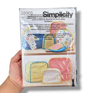 May include: A Simplicity sewing pattern packet featuring various quilted baby items. The packet includes images of bibs, burp cloths, and other accessories in yellow, pink, and blue patterns. The packet is held in a person's hand.
