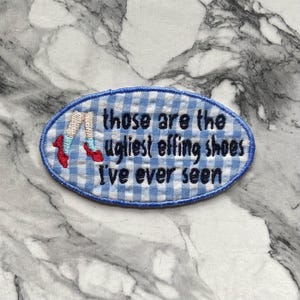 May include: An oval-shaped embroidered patch featuring a light blue and white gingham pattern. The patch displays an illustration of legs wearing light blue socks and red shoes, with the black embroidered text: those are the ugliest effing shoes I've ever seen.