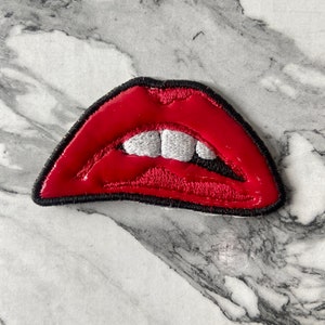 May include: An embroidered patch shaped like a pair of open lips. The lips are a vibrant glossy red with a thick black stitched border. Inside the mouth, two white teeth are visible against a dark background, creating a high-contrast design.