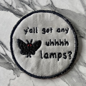 May include: A circular embroidered patch with a silver-grey background and a black border. It features a black moth with small red eyes and the black embroidered text: y'all got any uhhhh lamps? The patch has a subtle, repeating floral pattern.
