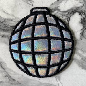 May include: A circular embroidered patch featuring a disco ball design. The patch has a black stitched grid pattern over a shimmering, iridescent silver fabric that reflects light with subtle rainbow hues, creating a multi-faceted appearance.