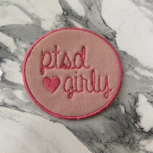 May include: A circular embroidered patch with a dusty rose background and a dark pink stitched border. The text "ptsd" is embroidered in dark pink cursive above a small dark pink heart, followed by the word "girly" in dark pink cursive script.