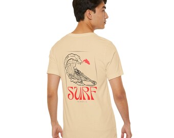 May include: A beige t-shirt with a black and white graphic of a skeleton surfing on a wave. The graphic is framed in a rectangle with the word "SURF" in red letters below it. The text "THINGS" is in smaller red letters below the word "SURF".