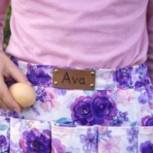 May include: A floral egg gathering apron with purple and white flowers. The apron has multiple pockets and a brown leather tag that says "Ava". A person is holding a beige egg.