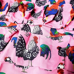 May include: Pink fabric with a repeating pattern of colorful roosters. The roosters feature a variety of designs, including black and white, and vibrant red, blue, and green. The fabric is ruffled, suggesting it may be part of a garment.