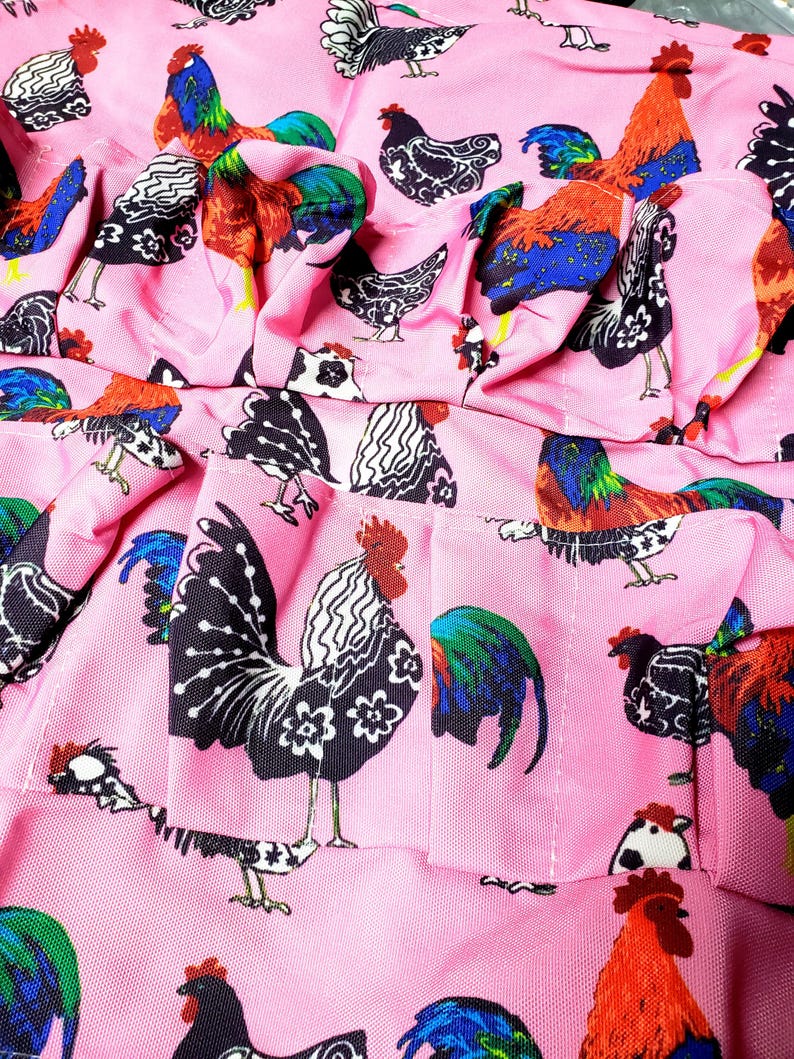 May include: Pink fabric with a repeating pattern of colorful roosters. The roosters feature a variety of designs, including black and white, and vibrant red, blue, and green. The fabric is ruffled, suggesting it may be part of a garment.