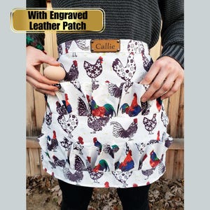 May include: White apron with a chicken and rooster print. The apron has multiple pockets and a ruffle trim. The text "With Engraved Leather Patch" is above the apron, and the name "Callie" is on a leather patch on the apron.