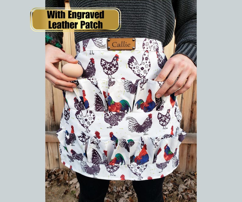 May include: White apron with a chicken and rooster print. The apron has multiple pockets and a ruffle trim. The text "With Engraved Leather Patch" is above the apron, and the name "Callie" is on a leather patch on the apron.