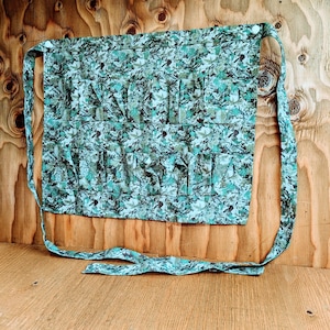 May include: A green and blue floral print apron with multiple pockets. The apron has long ties for securing around the waist and neck. The fabric has a pattern of leaves and other foliage.