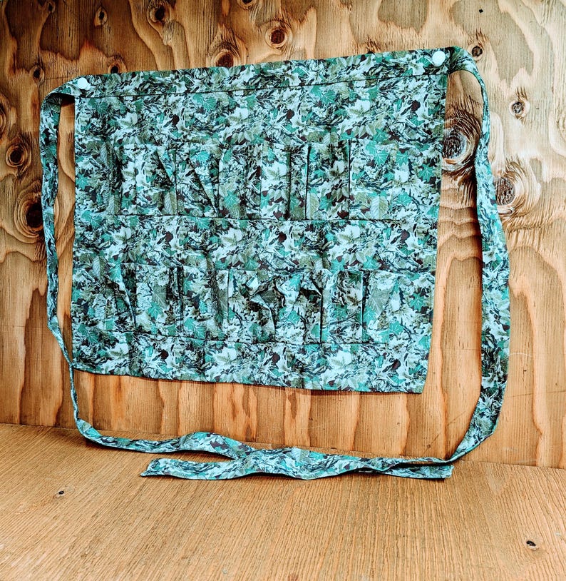 May include: A green and blue floral print apron with multiple pockets. The apron has long ties for securing around the waist and neck. The fabric has a pattern of leaves and other foliage.
