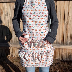 May include: White apron with a colorful rooster pattern. The apron has a pocket and ruffled egg-gathering pockets. The apron is worn by a person. The background is a wooden fence.