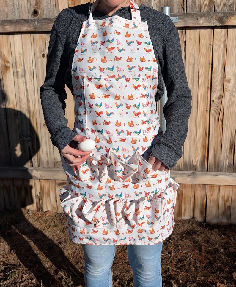 May include: White apron with a colorful rooster pattern. The apron has a pocket and ruffled egg-gathering pockets. The apron is worn by a person. The background is a wooden fence.