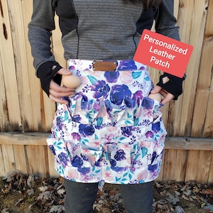 May include: A white apron with a floral print in shades of purple, pink, and blue. The apron has multiple pockets and a brown leather patch that says "Christy". A red sign says "Personalized Leather Patch".