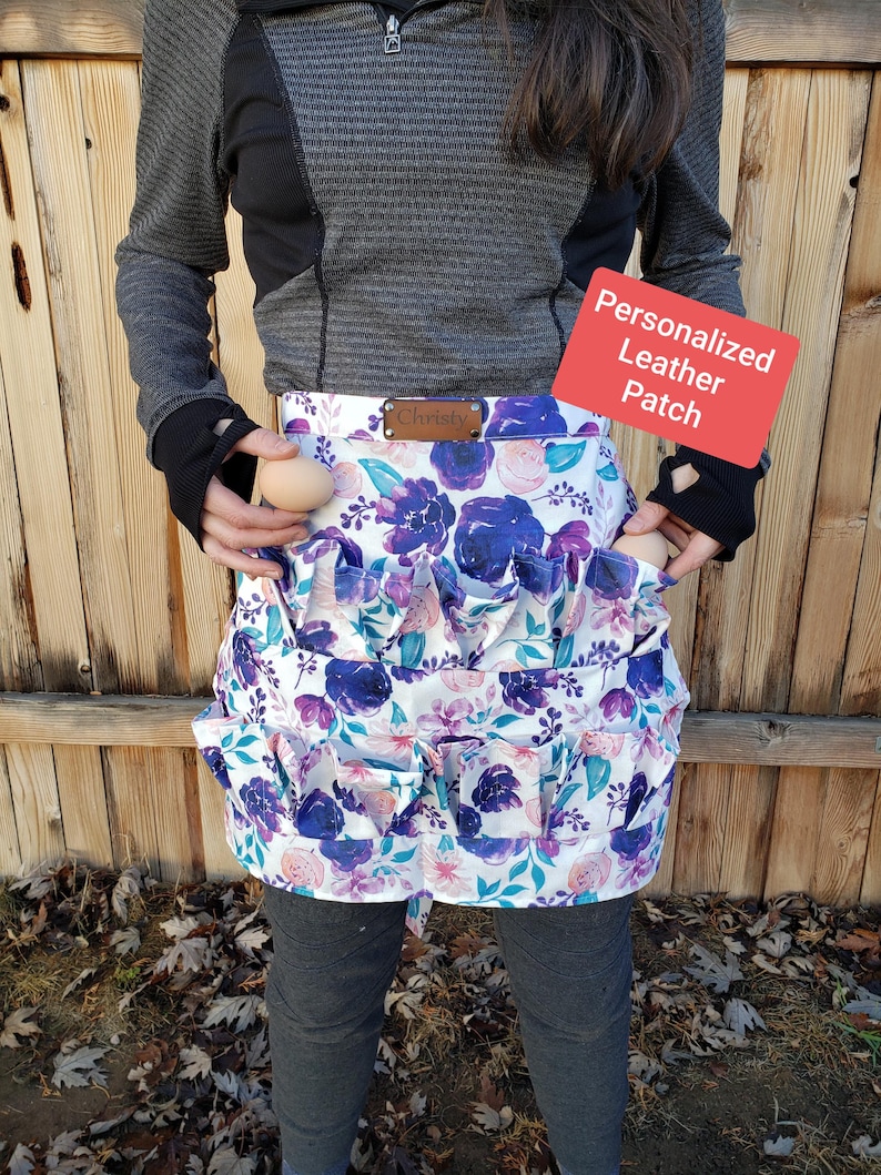 May include: A floral egg gathering apron with a white background and purple and blue flower pattern. The apron has multiple pockets for holding eggs. A small brown leather patch with the name "Christy" is sewn on the apron. The apron is designed for collecting eggs.