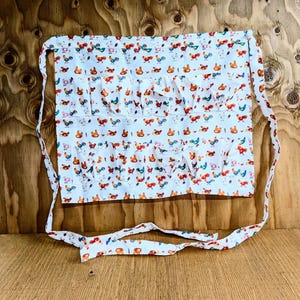 May include: White cotton apron with a colorful rooster print. The apron features multiple pockets and ruffled accents. The apron has long ties for securing around the waist. The rooster design includes red, blue, orange, and green accents.