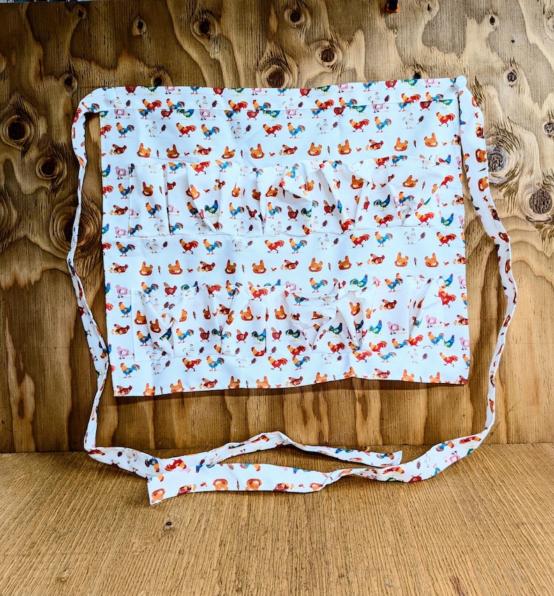 May include: White cotton apron with a colorful rooster print. The apron features multiple pockets and ruffled accents. The apron has long ties for securing around the waist. The rooster design includes red, blue, orange, and green accents.