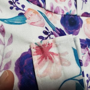 May include: Close-up of a white fabric with a floral pattern. The design features purple, pink, and teal flowers and leaves. The fabric has a textured appearance, and the edges are neatly sewn.