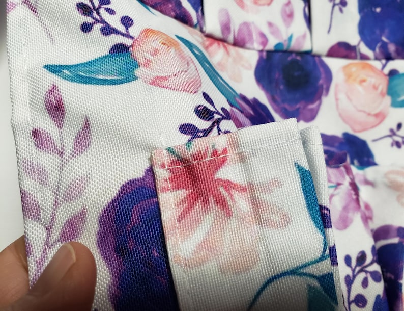 May include: Close-up of a white fabric with a floral pattern. The design features purple, pink, and teal flowers and leaves. The fabric has a textured appearance, and the edges are neatly sewn.