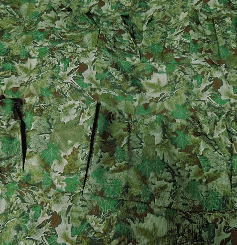 May include: Close-up of a fabric with a camouflage pattern. The design features a dense arrangement of green, brown, and white leaf shapes. The fabric appears to be pleated or gathered, creating a textured surface.