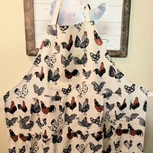 May include: White apron with a rooster and hen pattern. The apron has two pockets and a neck strap. The roosters are in various colors, including red, black, and white. The apron is hanging in front of a framed picture with the words "Country Chicks".