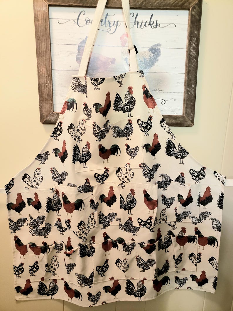 May include: White apron with a rooster and hen pattern. The apron has two pockets and a neck strap. The roosters are in various colors, including red, black, and white. The apron is hanging in front of a framed picture with the words "Country Chicks".