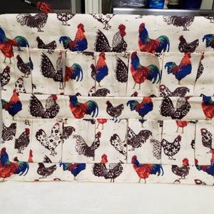 May include: Cream-colored fabric egg holder with a repeating pattern of roosters and hens in red, blue, and black. The holder has multiple pockets for storing eggs. The fabric is a light color, and the design is detailed.