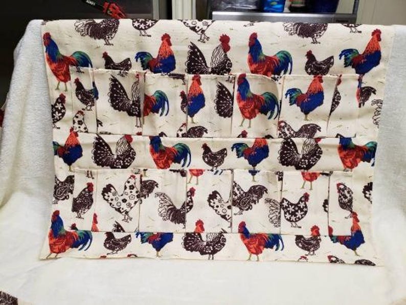 May include: Cream-colored fabric egg holder with a repeating pattern of roosters and hens in red, blue, and black. The holder has multiple pockets for storing eggs. The fabric is a light color, and the design is detailed.