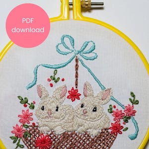 May include: Embroidered hoop art featuring two white bunnies sitting in a brown basket with pink and green flowers. A blue ribbon with a bow is above the basket.