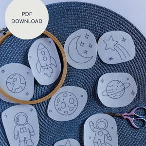 以下が含まれることがあります： ロケット、惑星、星、宇宙飛行士など、宇宙をテーマにした黒い線画が描かれた白い布パッチのコレクション。 パッチは青色のテクスチャ加工された表面に配置され、木製の刺繍枠とハサミが写っています。