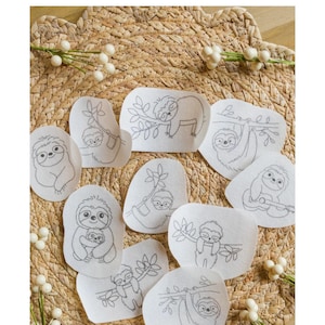 May include: Nine white felt patches with black line drawings of sloths on branches. The patches are arranged in a circle on a woven straw mat.