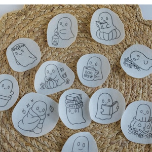 May include: A collection of white fabric patches with black embroidered ghost designs. The ghosts are depicted in various poses, some holding objects like books, lanterns, and flowers. The text "Stick and Stitch" is at the top.
