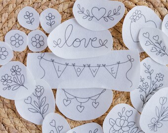 May include: A collection of white fabric patches with delicate black embroidery. Designs include flowers, hearts, and the word "love". Some patches feature floral arrangements, while others display simple heart and leaf motifs. The patches are arranged on a woven, light brown surface.