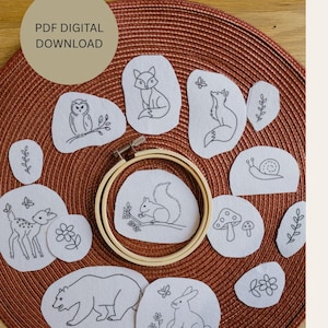 Könnte beinhalten: Eine Sammlung weißer Filzdesigns mit schwarzer Stickerei, darunter Tiere und Naturmotive wie Fuchs, Eule, Eichhörnchen, Bär, Reh, Hase, Schnecke, Pilze, Blumen und Blätter. Der Text "PDF DIGITAL DOWNLOAD" befindet sich in einem Kreis. Ein hölzerner Stickrahmen ist in der Mitte.