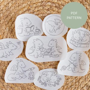 May include: A selection of white fabric patches featuring embroidered dragon designs. The dragons are shown in different scenarios, such as reading, having tea, and toasting marshmallows. The text "PDF PATTERN" is in a green circle.