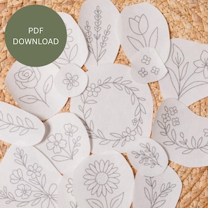 May include: A collection of white felt floral designs with gray embroidered outlines, including roses, tulips, and wreaths. The designs are scattered on a woven surface. A green circle with the text "PDF DOWNLOAD" is in the upper left corner.