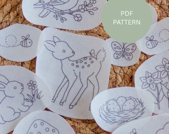 May include: A collection of white fabric patches with embroidered nature-themed designs, including a bird on a branch, a deer, a butterfly, bees, a rabbit, flowers, a mushroom, and a nest with eggs. The text "PDF PATTERN" is also visible.