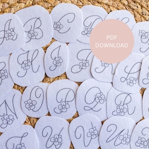 以下が含まれることがあります： 白いフェルトのアルファベット文字、花の模様、各文字は黒で縁取られています。文字は織り込まれた表面に配置されています。「PDF DOWNLOAD」というテキストが入ったピンク色の円が背景にあります。