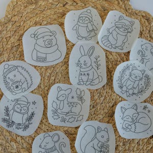 May include: A collection of white fabric patches with black line drawings of winter animals, including a bear, penguin, raccoon, rabbit, hedgehog, owl, deer, cat, fox, mouse, and squirrel. The patches are arranged on a woven, light brown surface.