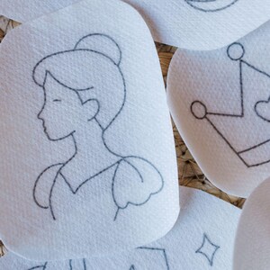 May include: Close-up of several white, oval-shaped patches with delicate black line drawings. One patch features a profile of a woman with her hair in a bun. Other patches show a crown and a diamond shape. The patches are arranged on a textured, woven surface.