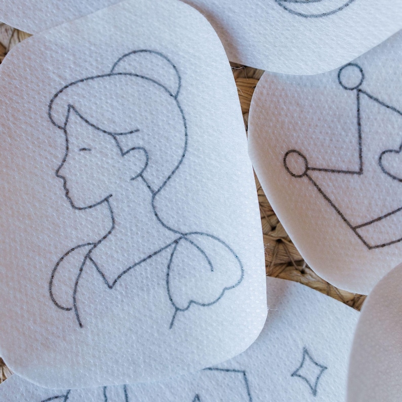 May include: Close-up of several white, oval-shaped patches with delicate black line drawings. One patch features a profile of a woman with her hair in a bun. Other patches show a crown and a diamond shape. The patches are arranged on a textured, woven surface.