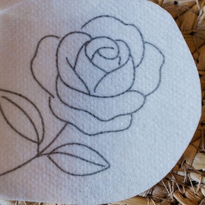 May include: A white, circular patch featuring a detailed, black outline of a rose. The rose is depicted in full bloom with multiple petals and a stem with two leaves. The design is centered on the patch.