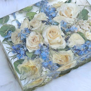 May include: A rectangular resin art piece featuring a bouquet of cream-colored roses and blue delphiniums. The flowers are preserved within the clear resin, with green foliage accents. The piece is set against a white, fluffy background.