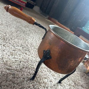 Puede incluir: Una olla de color cobre con un asa de madera y patas de metal negro. La olla tiene una textura martillada y un pequeño pico. El asa está unida con una pieza de metal retorcido. La olla está diseñada para cocinar.