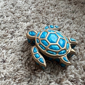 May include: A decorative turtle figurine with a gold-coloured frame and bright blue accents. The turtle's shell and body are detailed with a mosaic-like pattern. The figurine is resting on a textured, light-coloured surface.