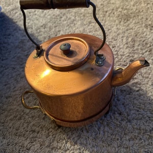 Peut inclure: Une bouilloire en cuivre avec une poignée en bois et une anse en métal incurvée. La bouilloire a un bec et un petit couvercle avec un bouton. La bouilloire repose sur une base en métal.