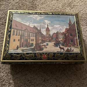 May include: A rectangular tin with a detailed illustration of a European town square. The tin has a gold-coloured frame with ornate blue and gold detailing. The illustration features buildings, people, and horse-drawn carriages.