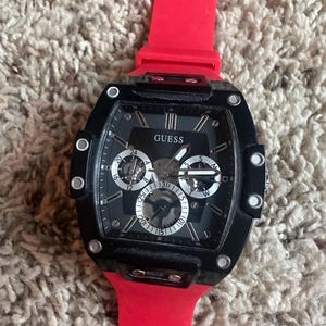 May include: A black Guess watch with a rectangular face and a red silicone strap. The watch face features multiple sub-dials and the brand name "GUESS". The watch is set against a textured, neutral-coloured background.